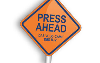 Ein Schild mit der Aufschrift PRESS AHEAD. Orangener Hintergrund und Blaue CI-Schrift der BJV