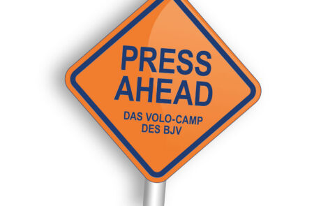 Ein Schild mit der Aufschrift PRESS AHEAD. Orangener Hintergrund und Blaue CI-Schrift der BJV